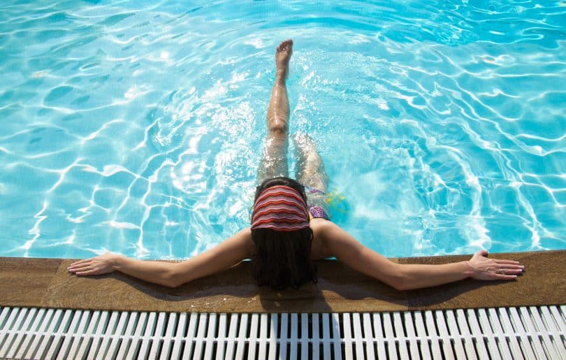 Imagem de uma piscina com uma água azul e limpa. Dentro dela uma mulher encostada na beirada relaxando as suas pernas.