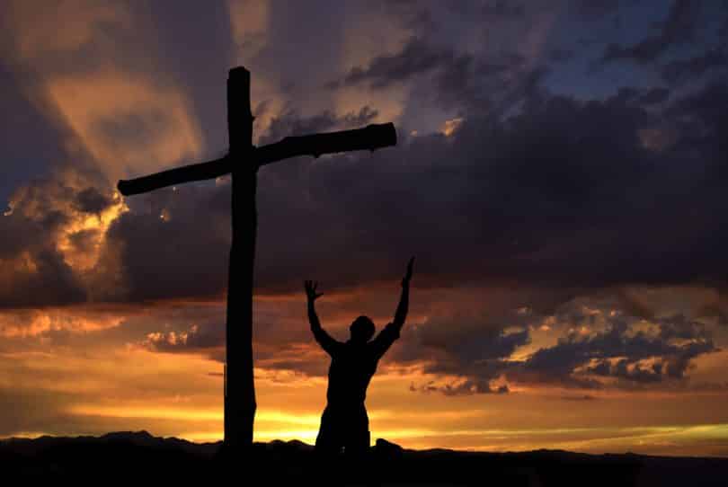 Silhueta de homem agachado próximo de uma cruz. Ao fundo, cenário de pôr do sol.