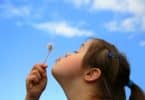 Imagem de uma criança portadora da síndrome de down. Ao fundo o céu azul e algumas nuvens brancas. A criança está feliz e assopra um dente de leão.