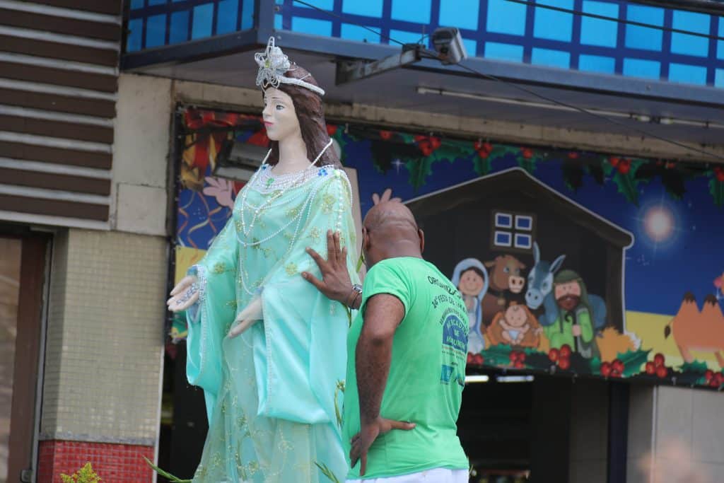 Imagem da estátua de Iemanjá no meio de uma rua. Ao lado dela, um filho de santo usando uma camiseta verde. Ele está com uma de suas mãos sobre ela.
