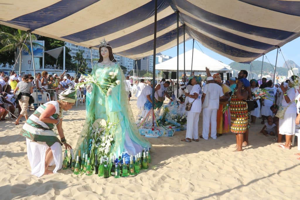 Imagem da estátua de Iemanjá embaixo de uma tenda na areia. Uma filha de santo faz homenagens à ela colocando varias garrafas de champanhe e flores brancas.