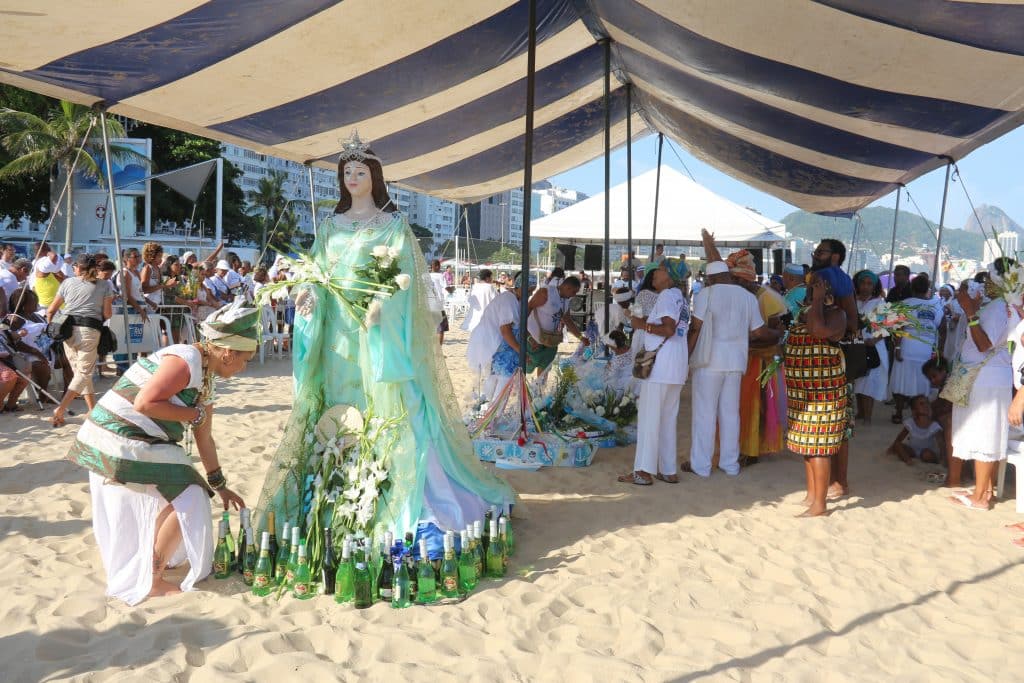 Imagem da estátua de Iemanjá embaixo de uma tenda na areia. Uma filha de santo faz homenagens à ela colocando varias garrafas de champanhe e flores brancas.
