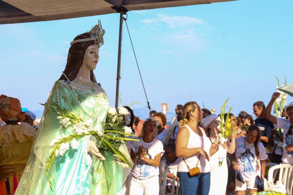 Imagem da estátua de Iemanjá na praia. Ao lado vários devotos dela cantando e fazendo oferendas como flores.
