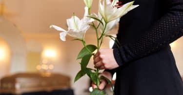 Imagem de uma mulher vestindo uma roupa preta dentro de uma sala de velório. Ela está segurando um ramo de flor branca e está ao lado do caixão.