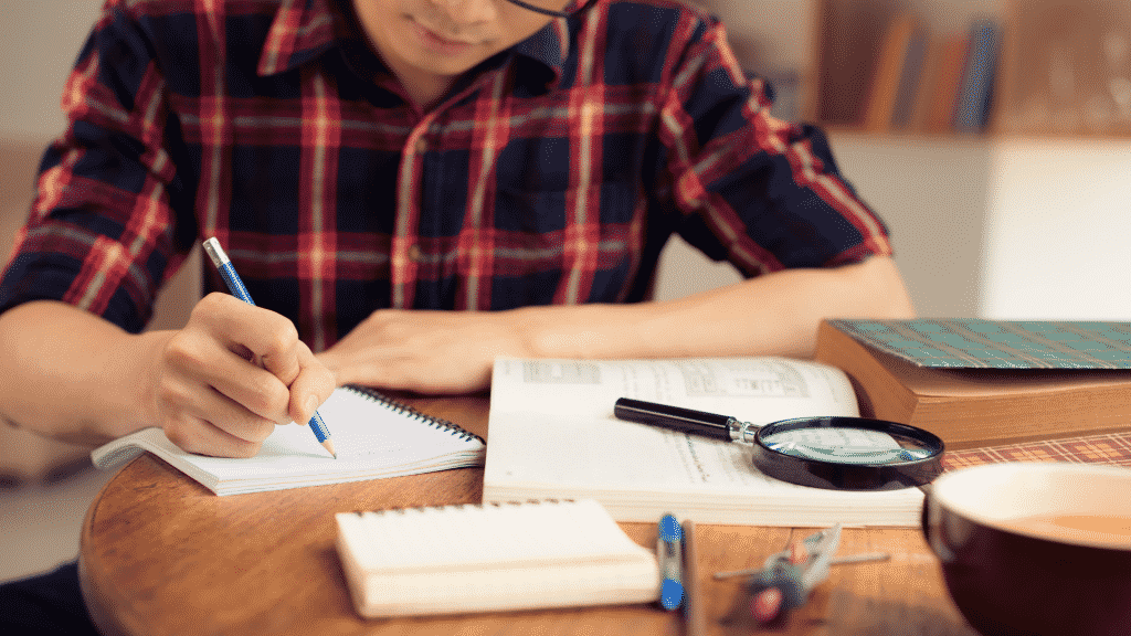 Garoto estudando e escrevendo anotações com livros em lupa sobre a mesa