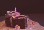 Imagem de um pedaço de brownie de chocolate disposto sobre uma travessa. Ele está decorado com um laço e um flor cor de rosa.