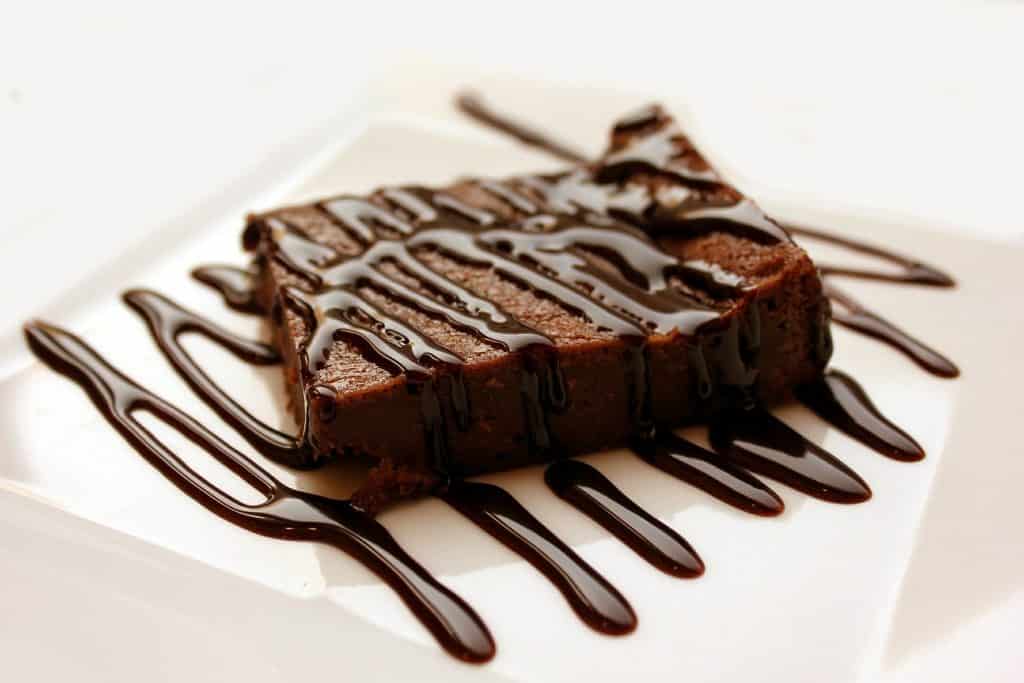 Imagem de um pedaço de brownie de chocolate vegano coberto com calda de chocolate, disposto em um prato branco de porcelana.