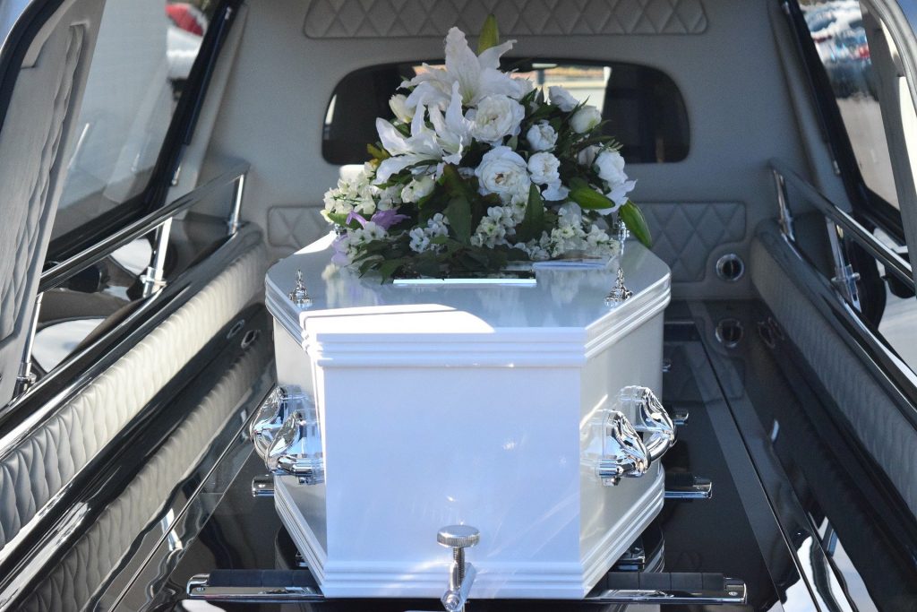 Imagem de um carro funeral e dentro dele um caixão branco com uma coroa de flores sobre ele. O carro está trazendo o corpo de uma pessoa para o velório.
