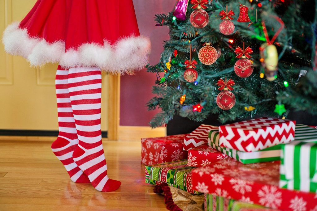 Imagem de uma mulher decorando a árvore de natal. Ela usa uma saia vermelha com pompom branco na barra e uma meia listrada nas cores vermelho e branco.