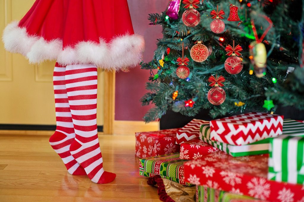 Imagem de uma mulher decorando a árvore de natal. Ela usa uma saia vermelha com pompom branco na barra e uma meia listrada nas cores vermelho e branco. 