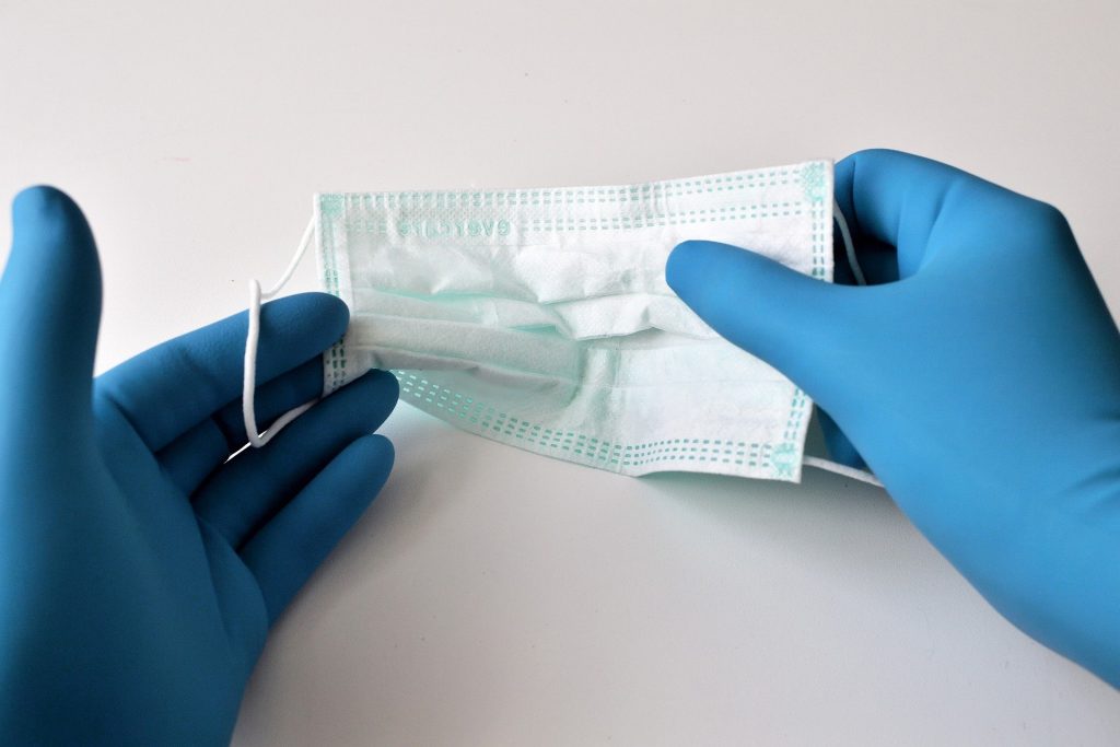 Imagem de duas mãos usando luvas emborrachadas de cor azul segurando entre os dedos uma máscara de proteção facial contra a Covid-19.