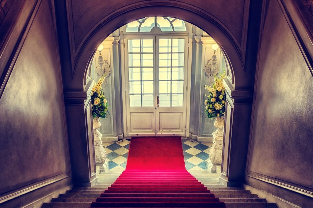 Imagem do hall de entrada de uma casa. Ao lado da porta dois vasos grandes de mármore com rosas-amarelas. Na porta até as escadas um tapete vermelho forra o piso.