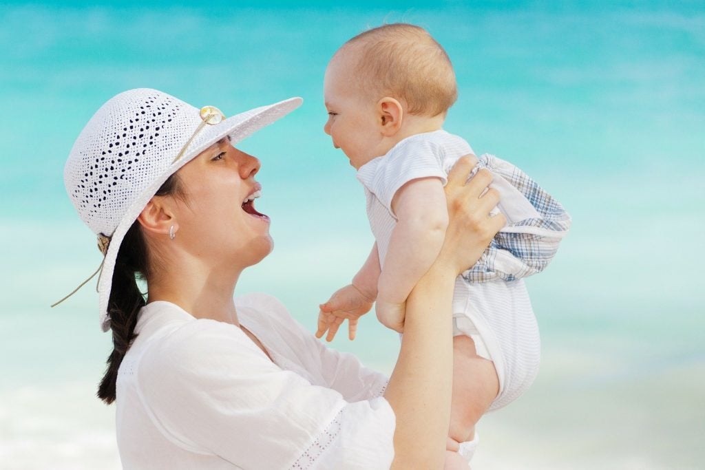 Imagem de uma jovem mãe segurando o seu bebê. Ela usa uma camisa e um chapéu de renda branco. O bebê usa um macacão branco. Ambos estão sorrindo.
