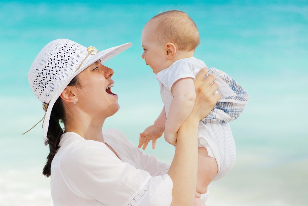 Imagem de uma jovem mãe segurando o seu bebê. Ela usa uma camisa e um chapéu de renda branco. O bebê usa um macacão branco. Ambos estão sorrindo.