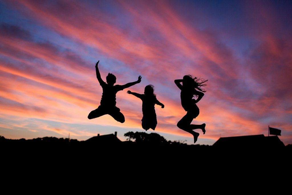 Imagem do céu e o por do sol à frente dele três pessoas amigas saltando.