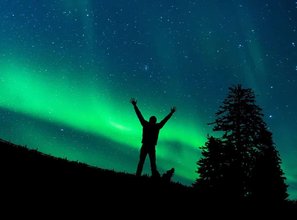 Silhueta de um homem com os braços abertos. Atrás dele está uma aurora boreal.