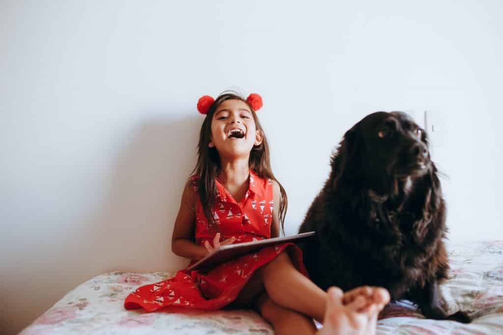 Menina ao lado de um cachorro preto. Ela está sorridente e segura um tablet nas mãos. 