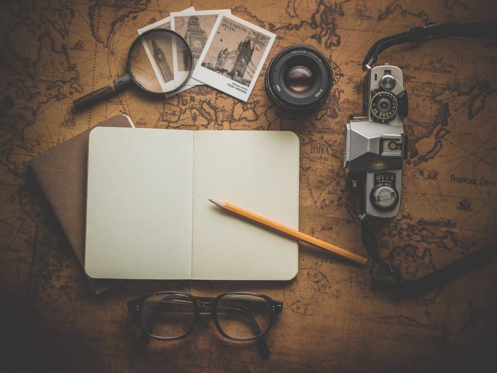 Um caderno em branco, fotografias, uma lupa, óculos e uma câmera fotográfica em cima de uma mesa de madeira   