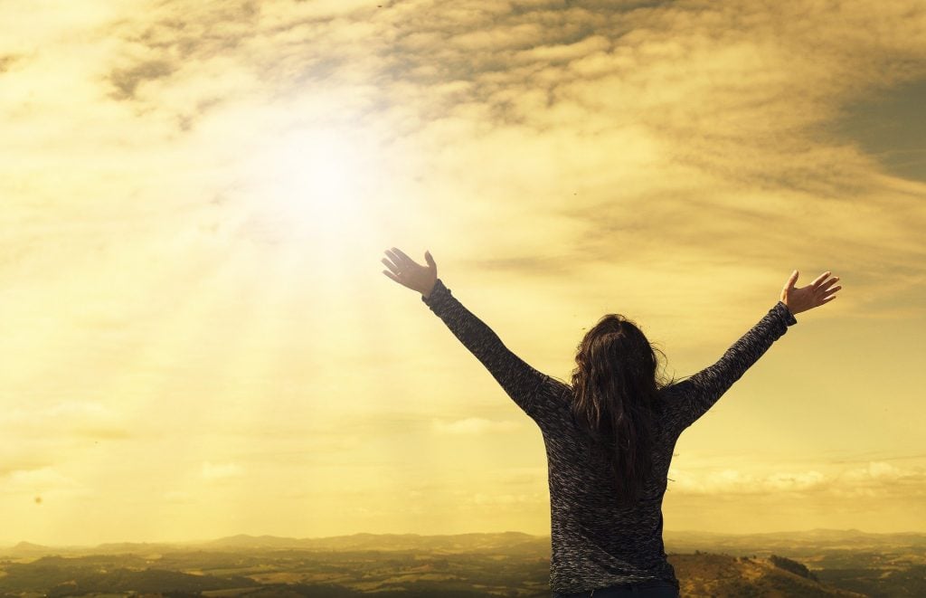 Imagem de uma mulher do alto de uma montanha. Ela está olhando para o sol de braços abertos, dando boas-vindas a ele.