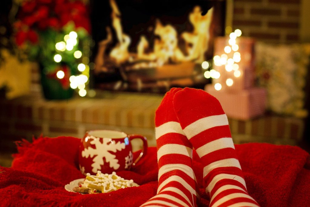 Imagem de um pé usando uma meia nas cores vermelho e branco. A pessoa está sentada no sofá com os pés elevados. Ela está sozinha passando o natal em isolamento. Ao lado um caneca vermelha e um pires com biscoitos natalinos.