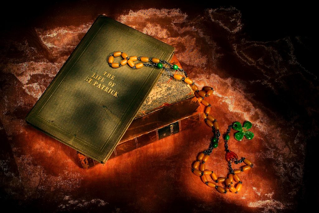 Imagem de uma bíblia e sobre ela um livro que traz a história de São Patrício. O livro tem capa dura na cor verde e ao lado um terço de contas e na ponta um trevo-de-quatro-folhas na cor verde.