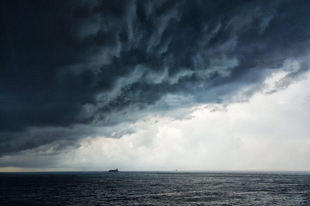 Imagem do oceano e sobre ele uma nuvem escura formando uma tempestade. Ao fundo um navio.
