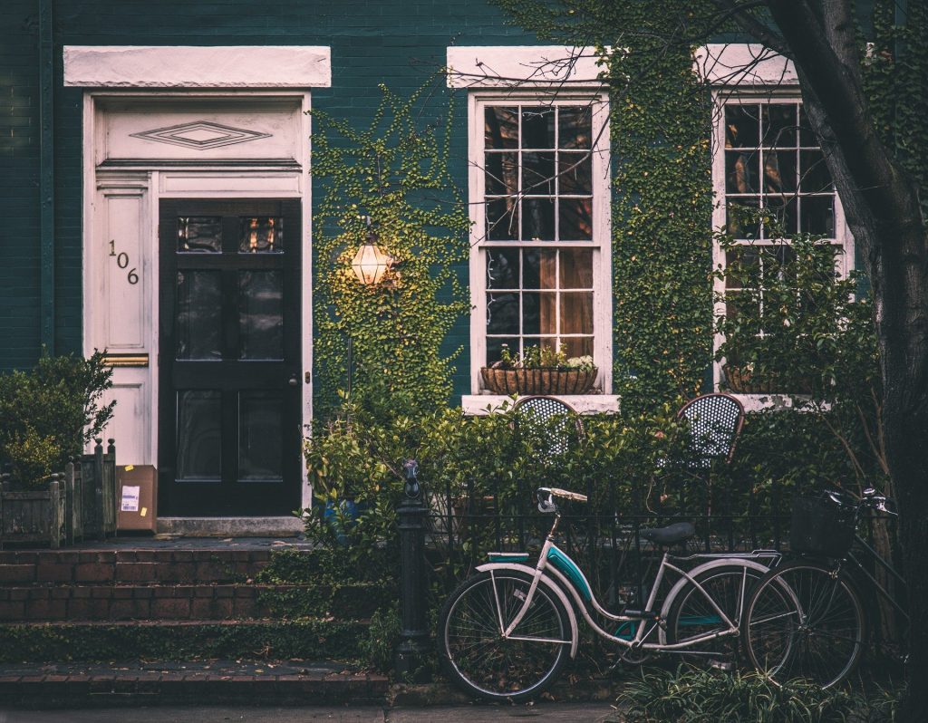 Imagem da frente de uma casa com muro coberto por uma folhagem. A porta da casa é pintada de preto e as janelas possuem um beiral na cor branco. Na frente da casa uma bicicleta de modelo vintage.
Free-Photos por Pixabay 