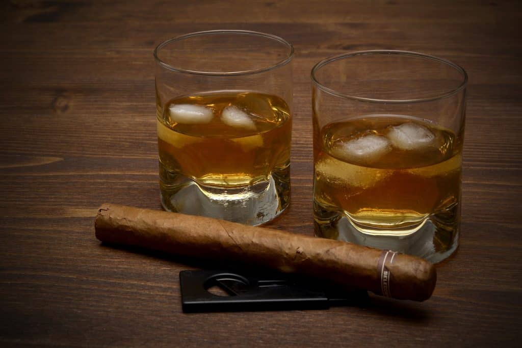 Imagem de dois copos com whisky e gelo e um charuto apagado. Eles estão dispostos sobre uma mesa de madeira.