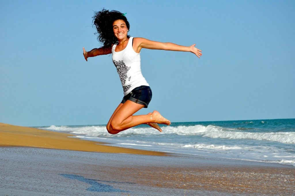 Imagem de uma mulher de cabelos escuros. Ela usa um short jeans e uma camiseta regata branca. Ela está dando um salta de braços abertos na areia da praia.