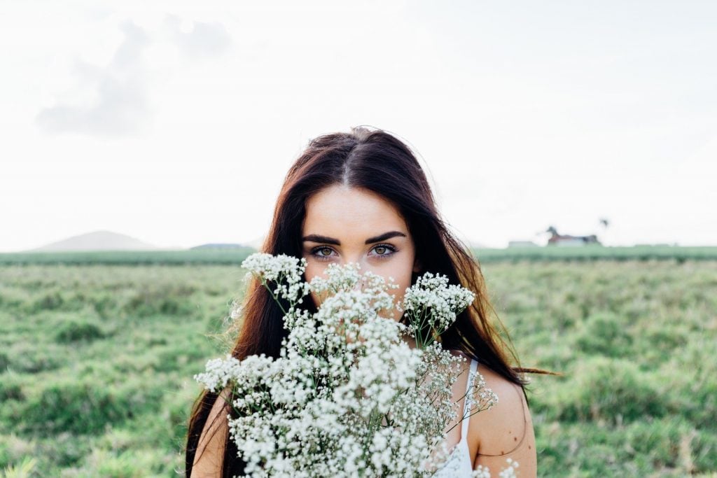 Imagem de uma bela mulher de cabelos longos e escuros em um campo. Ela segura em suas mãos um buquê de flores brancas.