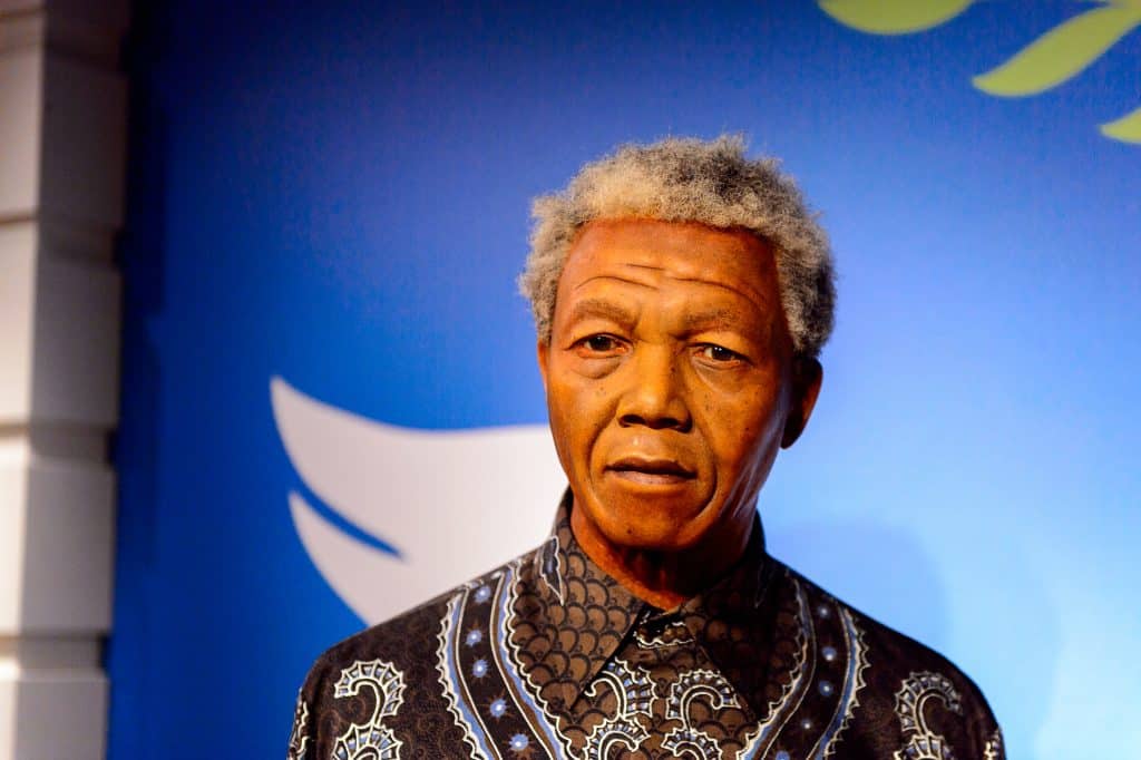 Imagem com fundo azul e a frente a foto de Nelson Mandela. Ele está usando uma camisa marrom com detalhes em branco e azul.