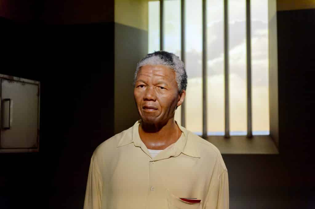 Imagem de Nelson Mandela na cela da prisão. Ele usa uma camisa bege e por baixo dela  uma camiseta de malha na cor branca.
