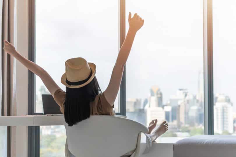 Mulher sentada em uma cadeira giratória observa cidade do alto de um prédio. Seus braços estão erguidos e ela usa um chapéu.