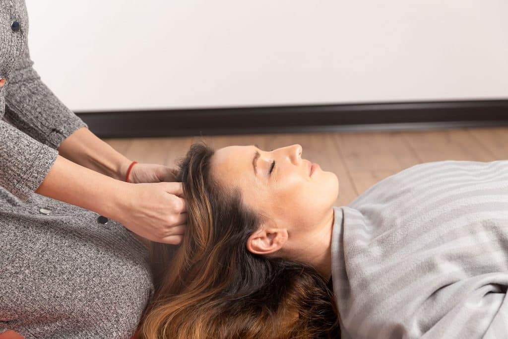 Mulher branca deitada de olhos fechados recebendo massagem na cabeça.