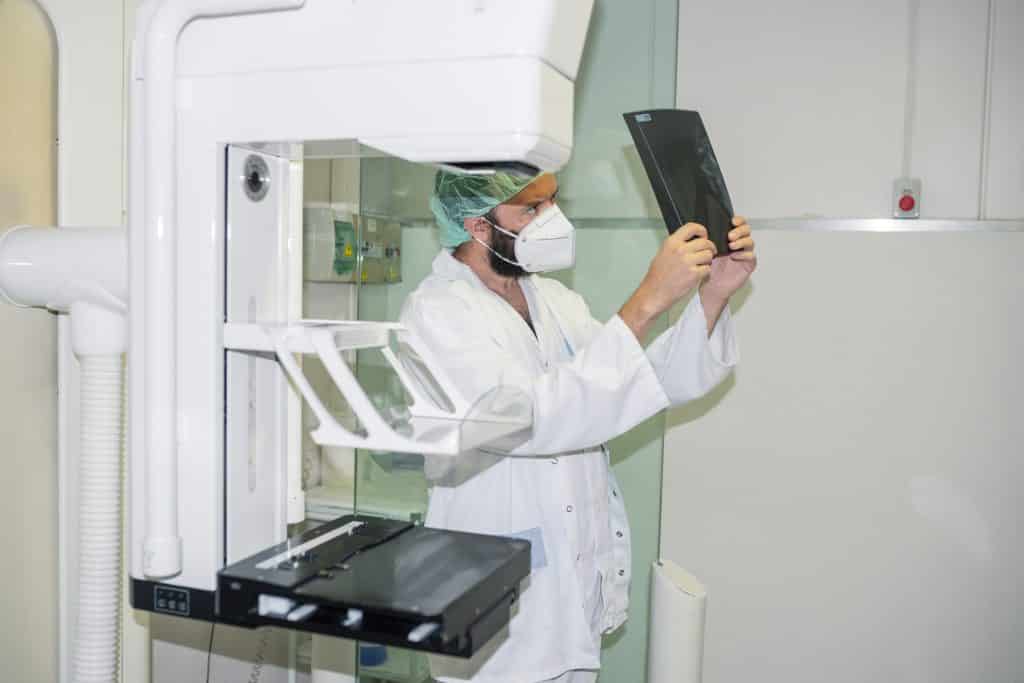 Imagem do equipamento de fazer mamografia e ao lado dele o médico examinando o resultado da paciente.
