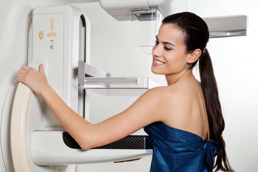 Imagem de uma paciente de cabelos longos ao lado da máquina de raio x. Ela está pronta para realizar o exame de mamografia.
