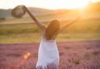 Mulher jovem com um vestido branco e um chapéu de palha em pé no meio de um campo com o pôr do sol ao fundo.