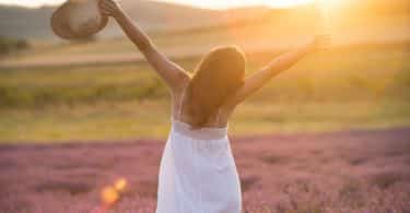 Mulher jovem com um vestido branco e um chapéu de palha em pé no meio de um campo com o pôr do sol ao fundo.