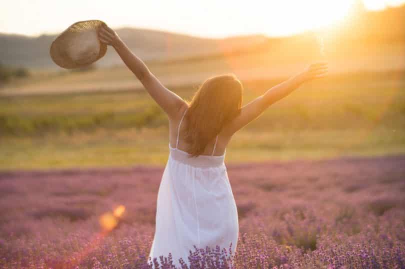 Mulher jovem com um vestido branco e um chapéu de palha em pé no meio de um campo com o pôr do sol ao fundo.