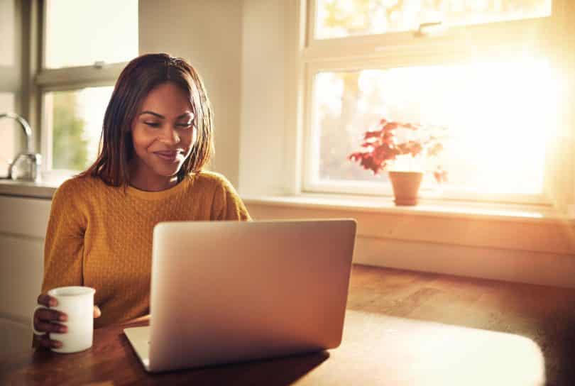 Mulher segura uma caneca de café e mexe em computador.