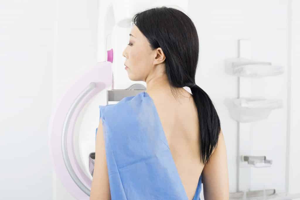 Imagem de uma mulher de costas com um tecido azul sobre um dos seus ombros. Ela está pronta para realizar a mamografia.

