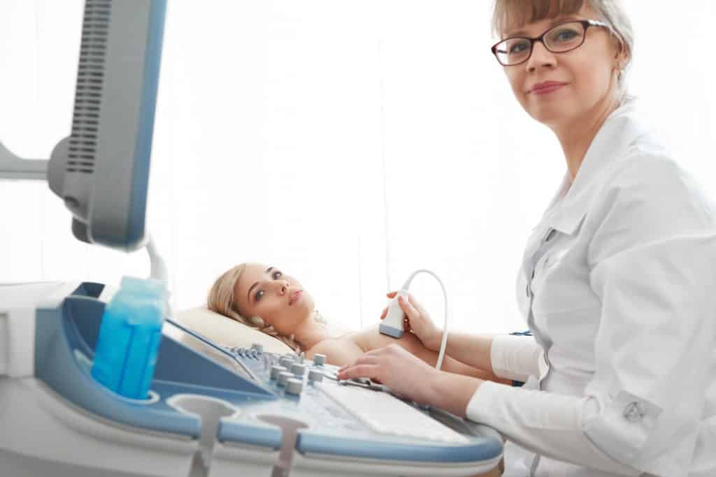 Imagem de uma paciente deitada sobre uma maca. Ela está sendo examinada pela sua médica e está fazendo uma mamografia.
