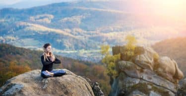 Mulher branca meditando sentada numa rocha.