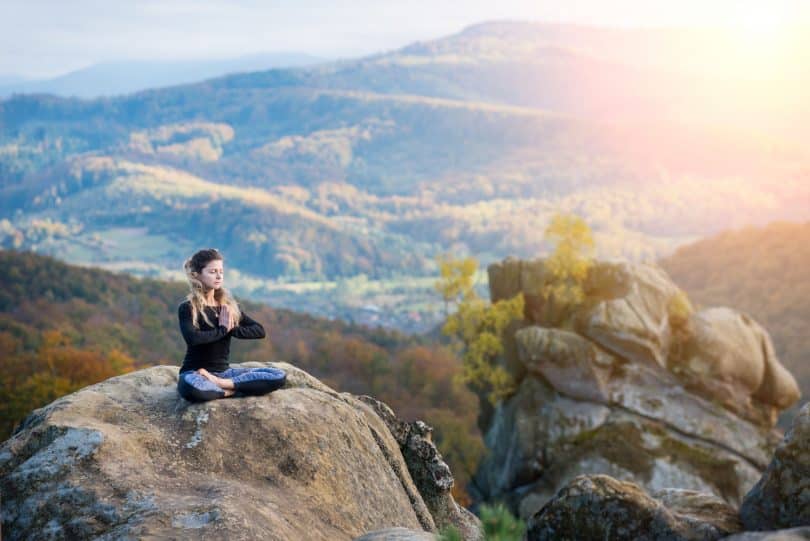 Mulher branca meditando sentada numa rocha.