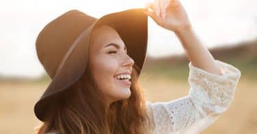 Mulher branca com expressão sorridente segurando um chapéu na cabeça.