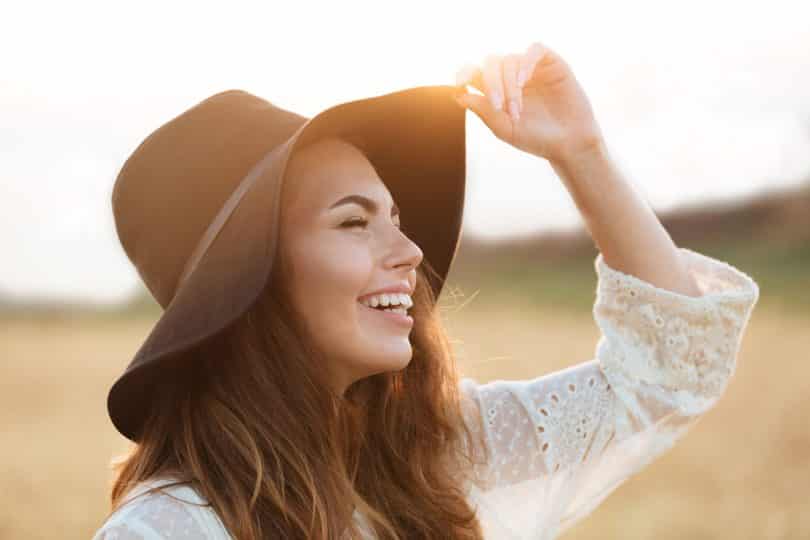 Mulher branca com expressão sorridente segurando um chapéu na cabeça.