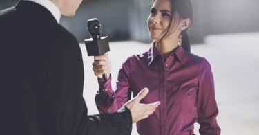 Um jornalista com uma camisa bordô sorrindo ouve um homem.