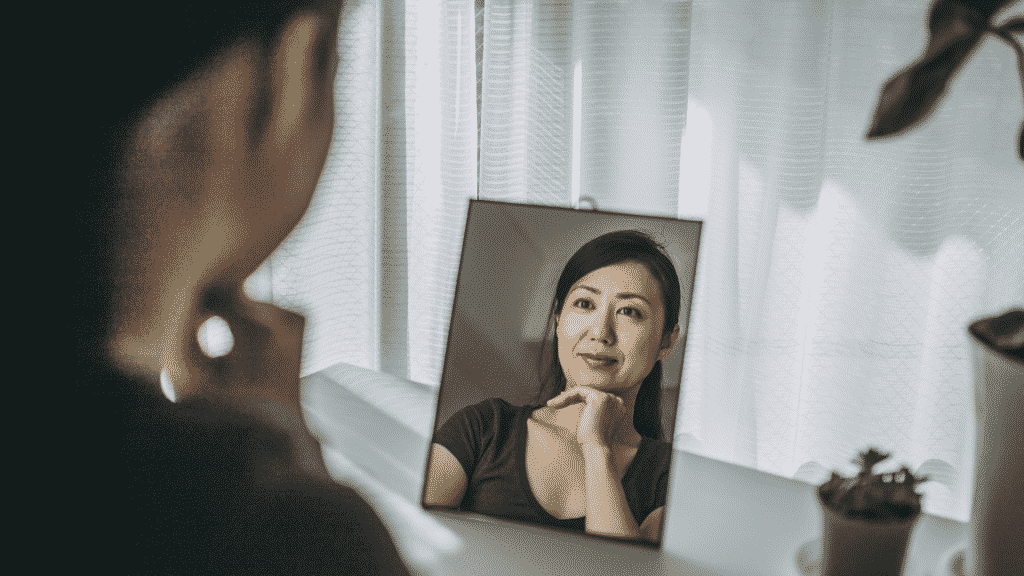 Mulher sorrindo enquanto analisa seu rosto no espelho.