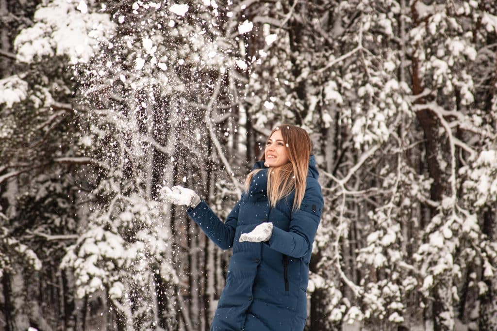 Imagem de uma floresta com suas árvores cobertas com neve. Em destaque temos uma mulher de cabelos claros e longos usando luvas e um casaco térmico na cor azul.