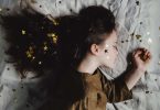 Imagem de uma linda garotinha usando um vestido marrom. Ela está dormindo em uma cama forrada com um lençol branco e sobre ela várias estrelas douradas.